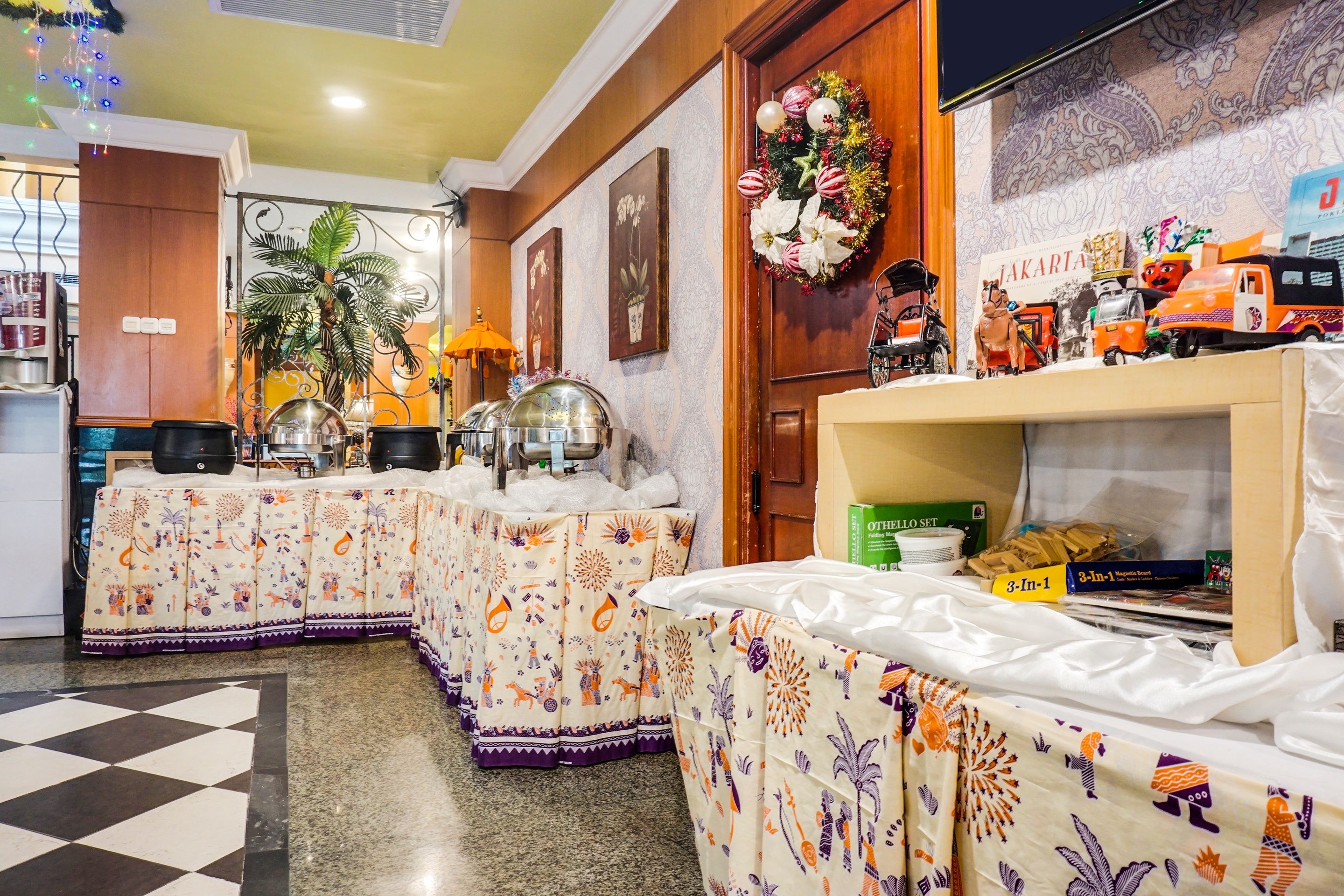 Une chambre d’hôtel avec un sol à carreaux, une table de buffet avec des plats couverts, un mur décoratif orné d’une guirlande, et un téléviseur fixé au mur.