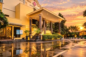 Entrada de hotel com varanda coberta, colunas e um espelho d'água refletivo.