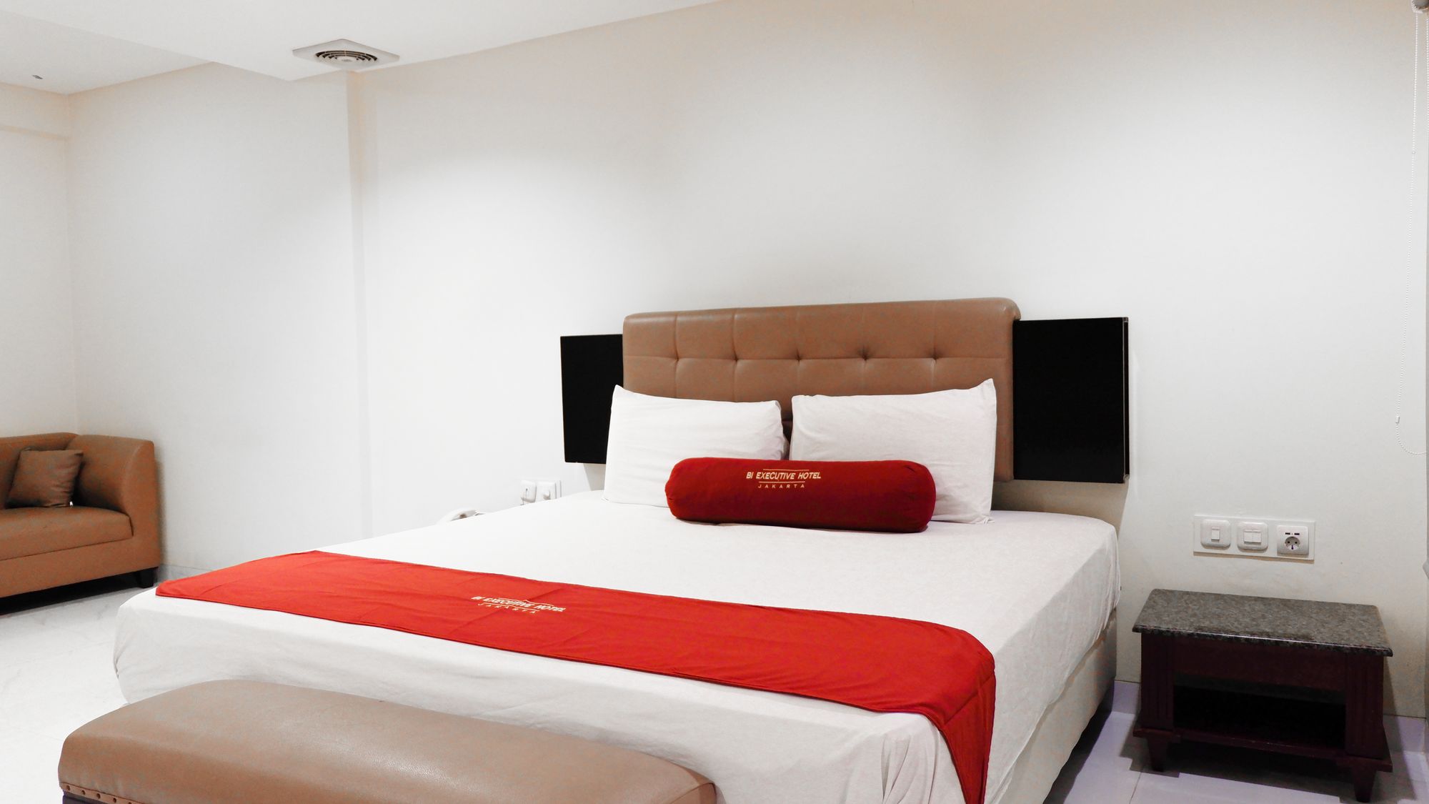 A hotel room with a bed, a red throw pillow, a brown headboard, a small bedside table, and a brown sofa.