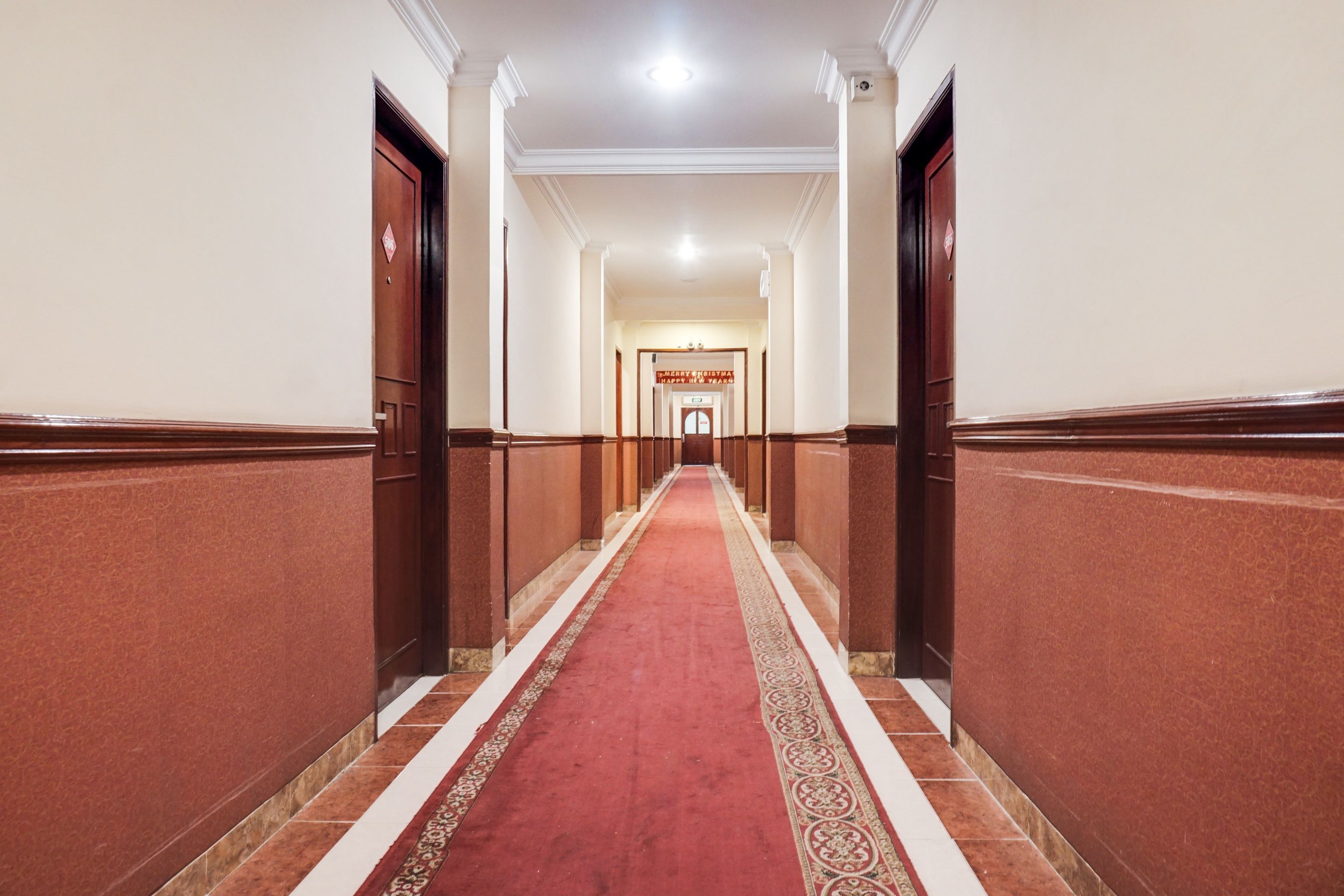 A hotel hallway with a red carpet, wooden doors, and a sign reading 'Merry Christmas.'