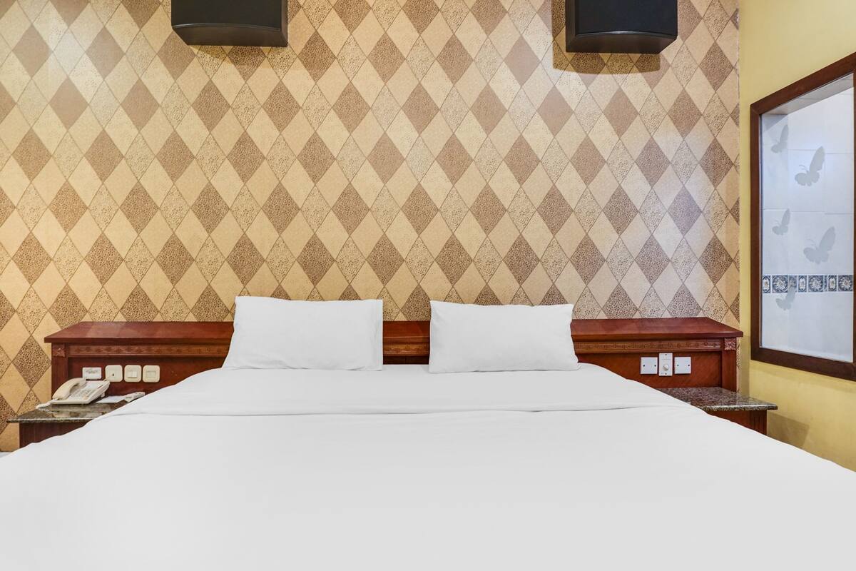 A hotel room with a large bed, two pillows, a wooden nightstand with a phone, and a wall with a diamond-patterned wallpaper.