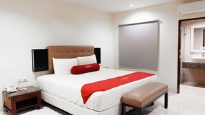 Una habitación de hotel con cama, una mesita de noche con teléfono, almohada roja, un banco, un ventanal grande y un baño visible a través de una puerta abierta.