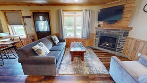 A cozy living room with a stone fireplace, a television mounted above it, a sofa with patterned cushions, and a dining area with a wooden table and chairs. - PA Grand Canyon, Mountain View, Pine Creek (Gaines)