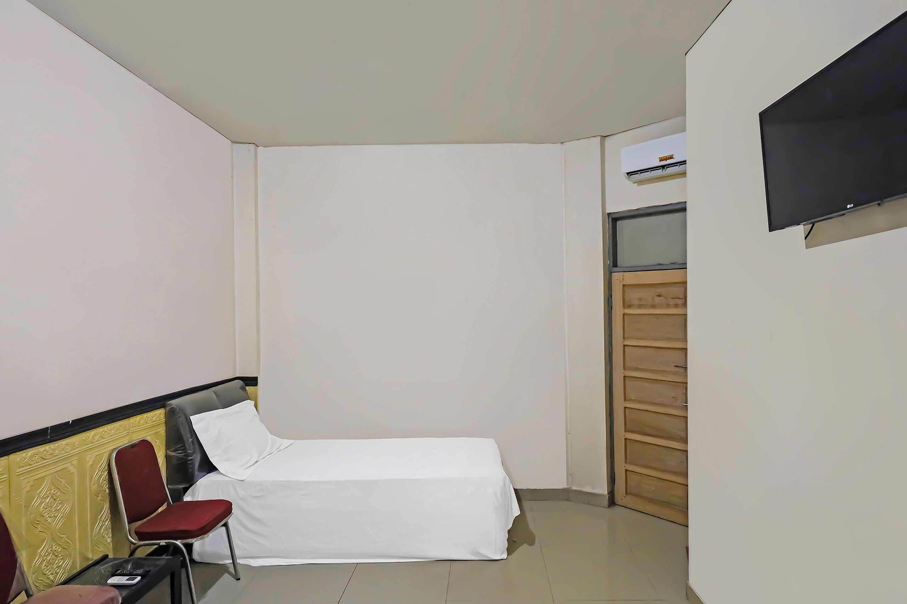 Une petite chambre avec un lit simple, une chaise rouge, un téléviseur fixé au mur et une porte en bois.