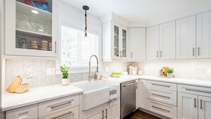 A modern kitchen with white cabinetry, a herringbone tile backsplash, a stainless steel dishwasher, and a window with blinds. - Harborside by AvantStay | Expansive Destin Retreat (Destin)