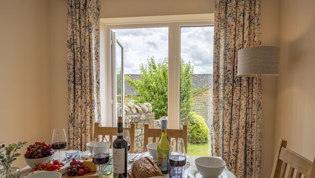 Una mesa de comedor preparada con diversos alimentos y bebidas, incluyendo uvas, queso y vino, con vista a un jardín a través de una ventana.