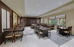 A spacious dining area with multiple tables and chairs, a marble floor, and large windows offering a view of greenery. - The Maharana Bagh (Udaipur)