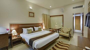 Habitación de hotel con una cama grande, mesitas de noche, un escritorio, una silla, un televisor, una ventana con cortinas y un ventilador de pared.