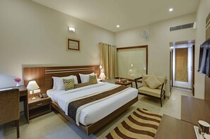Habitación de hotel con una cama grande, mesitas de noche, un escritorio, una silla, un televisor, una ventana con cortinas y un ventilador de pared.