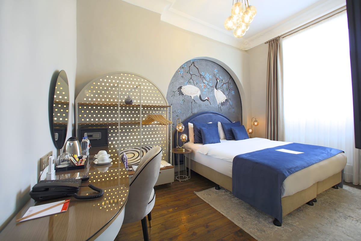 A hotel room with a bed, a desk, a chair, a mirror, and a wall with a decorative painting.