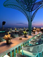 Un bar en la azotea con mesas al aire libre, plantas en macetas y vistas panorámicas de la ciudad al atardecer.