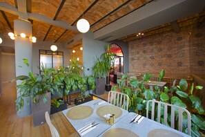 L’intérieur d’un restaurant présente des murs en briques, un plafond en bois et des plantes vertes. Les tables sont dressées avec des nappes blanches et sont mises avec soin.