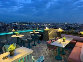 Una terraza en la azotea con varias mesas y sillas, plantas en macetas y vistas a la ciudad al anochecer.