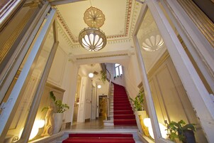 Una imponente escalera con alfombra roja que conduce a una plataforma, flanqueada por espejos ornamentados y plantas en macetas.