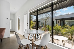 Un intérieur moderne avec une table de salle à manger ronde et quatre chaises, de grandes fenêtres donnant sur l’extérieur et une porte menant à une autre pièce.