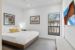 Une chambre à coucher comprenant un lit, une table de chevet, une lampe, des œuvres d’art encadrées et une fenêtre donnant sur des arbres et un immeuble.