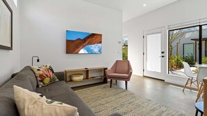 A modern living room with a grey sofa, a pink armchair, and a large window with a view of a garden. - Modern 1-bedroom Apartment - 80 Walkscore! (Phoenix)