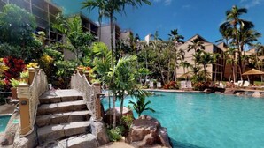 A pool area with clear blue water, surrounded by palm trees and a stone staircase leading to a building.