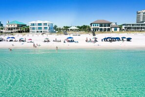 A beach with clear turquoise water, beach umbrellas, and several buildings in the background.