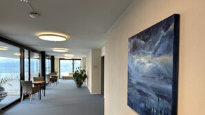 Un couloir d’hôtel moderne, orné d’un grand tableau abstrait au mur, avec un espace détente comprenant des tables et des chaises, et une vue dégagée grâce à de grandes fenêtres.