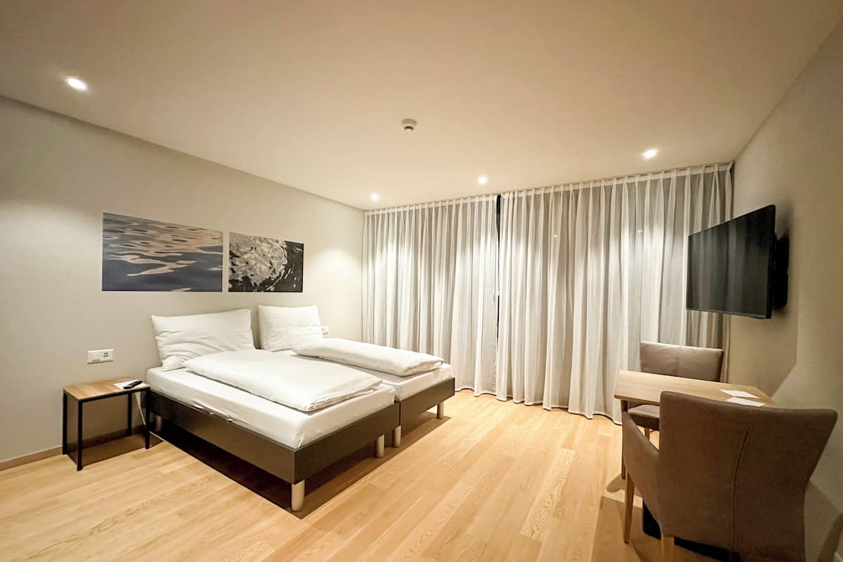 Una habitación de hotel moderna con una cama grande, un escritorio con silla, un televisor de pantalla plana y un amplio ventanal con cortinas.