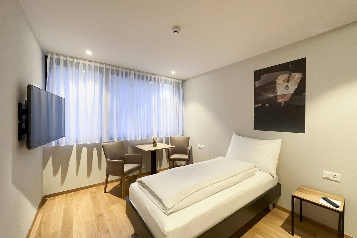 Habitación de hotel con cama, televisor montado en la pared, una mesita, dos sillas y una ventana con cortinas.
