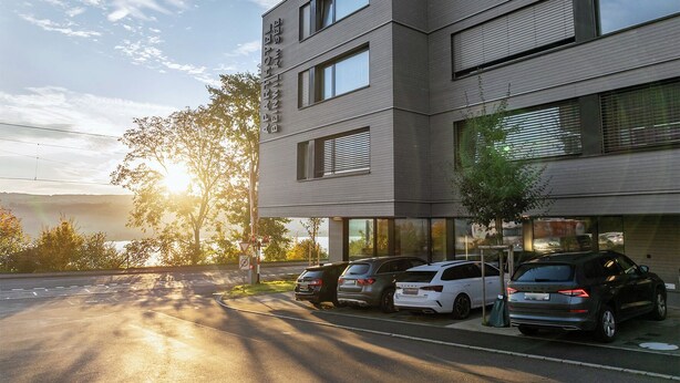 Un bâtiment moderne à plusieurs étages, doté de grandes fenêtres et d’un parking où sont stationnées plusieurs voitures.