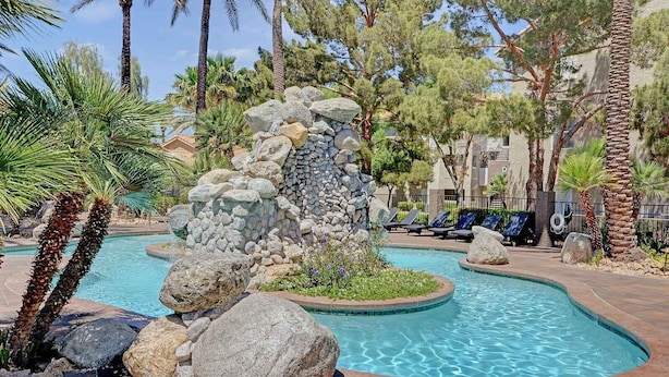 A pool area with a rock waterfall feature, lounge chairs, and palm trees. - Dreamy Vegas Strip area condo (Las Vegas)