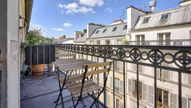 A balcony with a wooden table and chairs, a metal railing, and a view of adjacent buildings with dormer windows and chimneys. - Magnificent apartment ideally located in Paris (Paris)