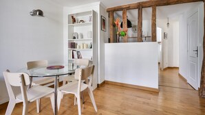 A dining area with a glass table and white chairs, a white bookshelf with various items, and a wooden beam structure. - Magnificent apartment ideally located in Paris (Paris)