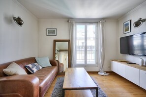 Uma sala de estar com sofá de couro marrom, mesa de centro de madeira, televisão de tela plana e uma janela grande com cortinas.