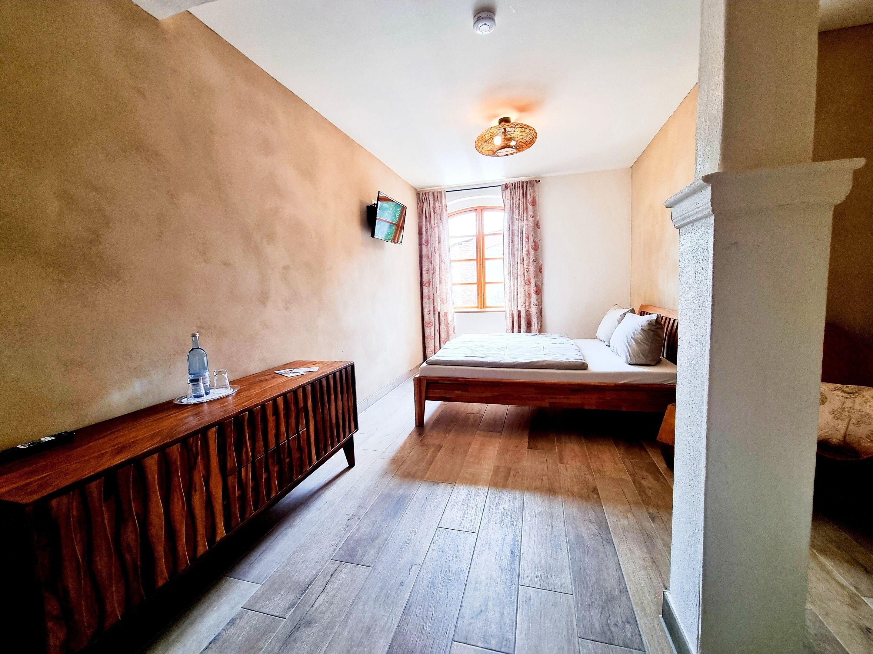 A bedroom with a wooden bed, a bedside table, a television, and a window with curtains.