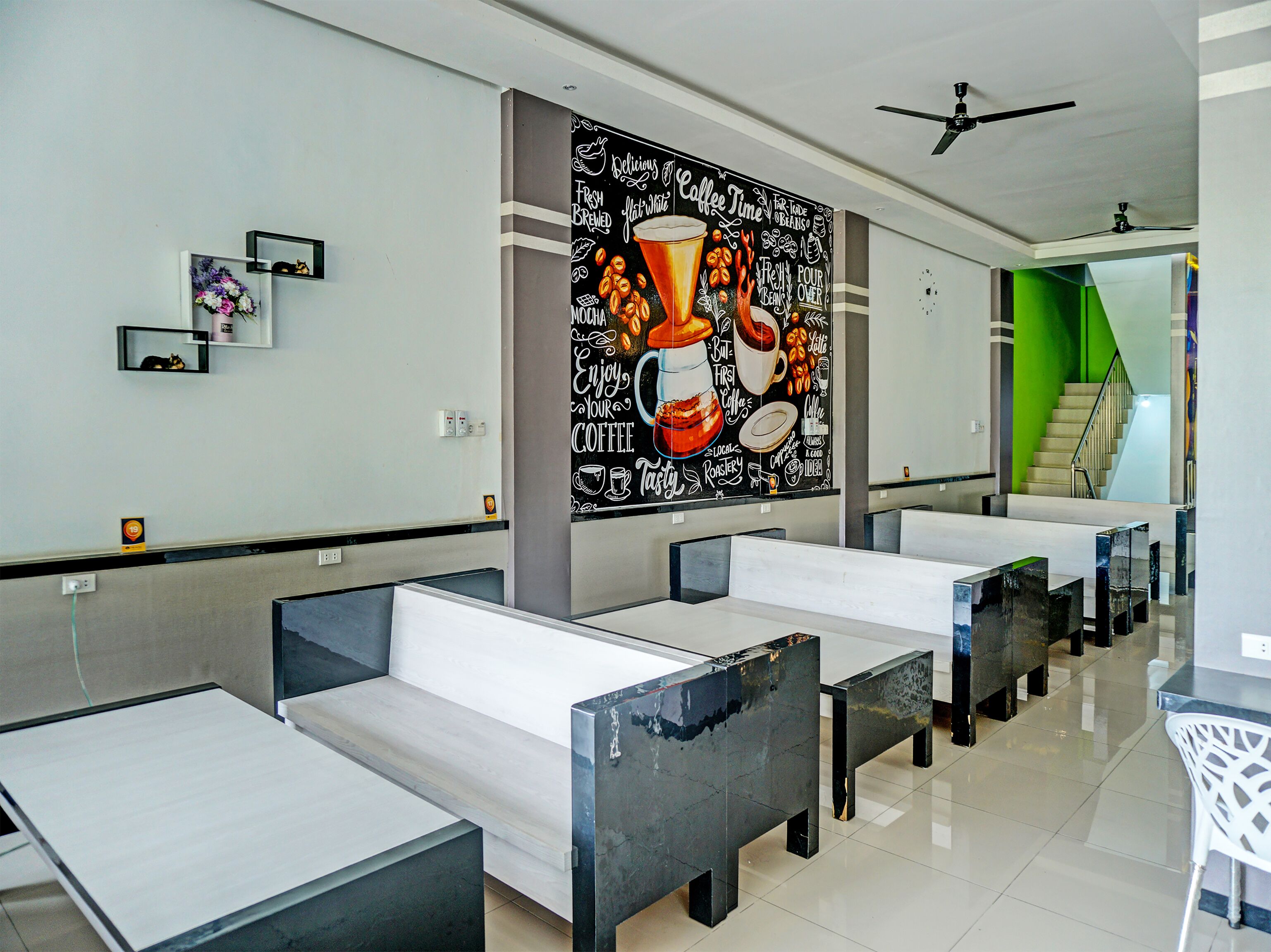 A modern cafe with a large coffee-themed mural, minimalist seating, and a staircase leading to a green area.