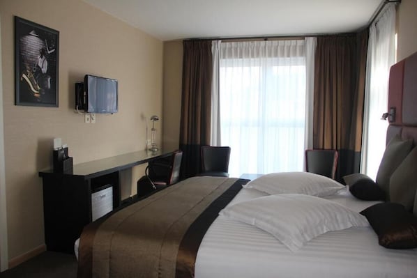 Habitación de hotel con cama, escritorio, silla, televisor y ventana con cortinas.