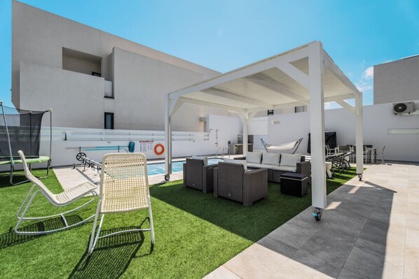 Un área de piscina en la azotea con sillones de descanso, un espacio de estar cubierto y un edificio moderno al fondo.