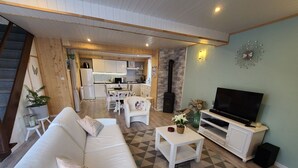 A living room with a sofa, a coffee table, and a TV. There is a kitchen in the background with white cabinets and a dining table. - Charming Cottage in Taintrux: Hiking, Skiing, Close to Saint-Dié & Alsace Attractions (Taintrux)
