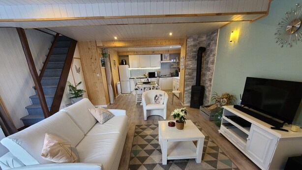 A modern living room with a sofa, coffee table, and TV stand. A staircase is visible in the background leading to a kitchen area. - Charming Cottage in Taintrux: Hiking, Skiing, Close to Saint-Dié & Alsace Attractions (Taintrux)