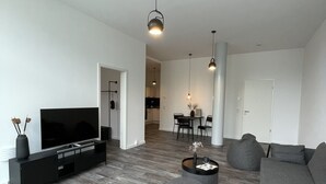 Ein modernes Wohnzimmer mit einem Flachbildfernseher, grauen Sofas und Holzboden. Im Hintergrund befinden sich zwei runde Couchtische und ein Essbereich.