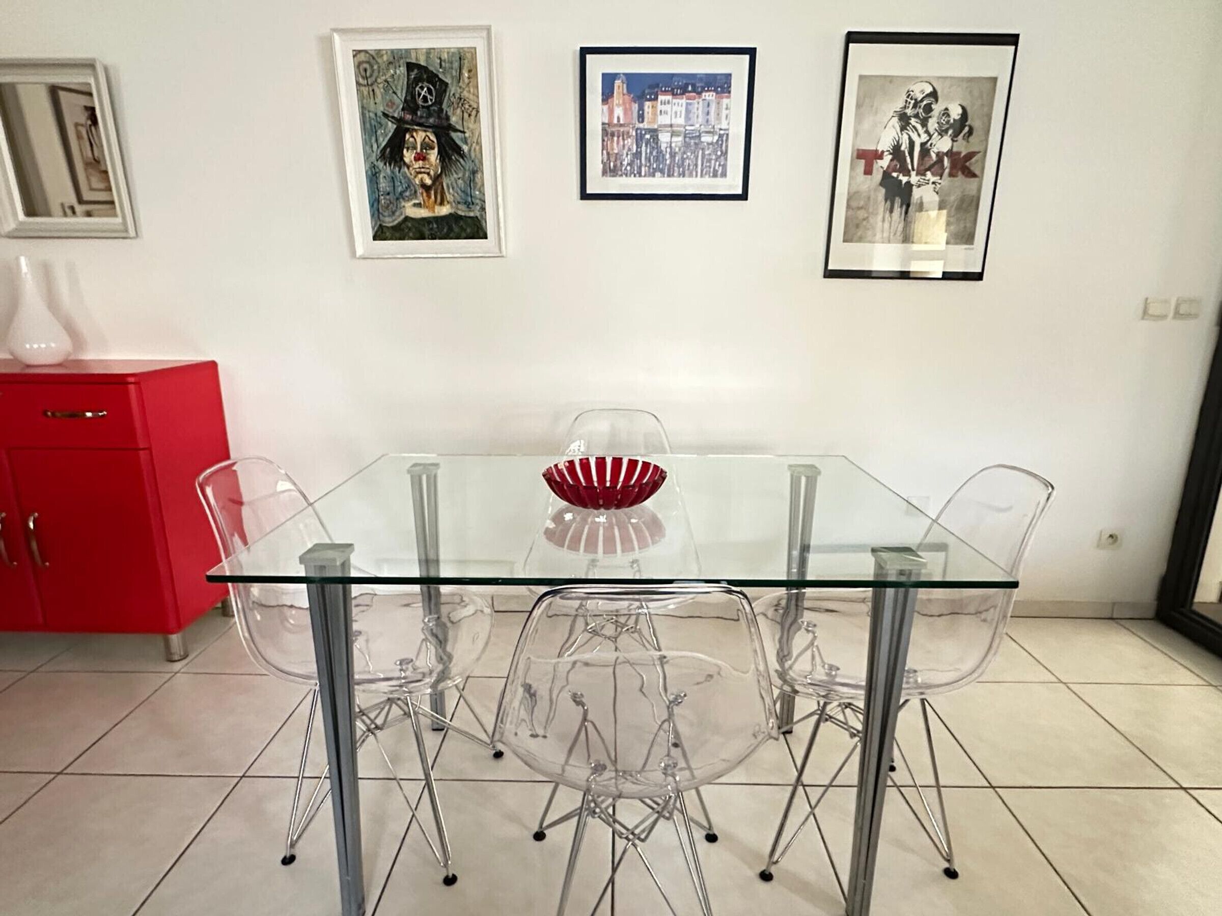 A dining area with a glass table and chairs, a red cabinet, and three framed artworks on the wall.