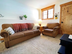 Een woonkamer met een bruine bank, een bruine fauteuil, een kleine tafel, een lamp en een houten deur.