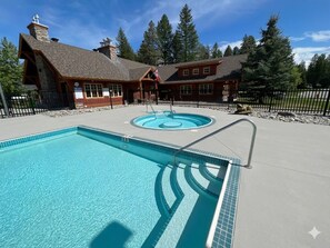 Un espace piscine comprenant une grande piscine rectangulaire et une plus petite piscine circulaire, le tout entouré d’un bâtiment en bois de style cabane, doté d’une cheminée en pierre.