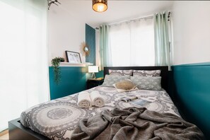 Un letto rifatto con cura, lenzuola decorate, una lampada da comodino e una piccola pianta su uno scaffale.