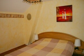 A bedroom with a wooden headboard, a checkered bedspread, a painting of two figures on a wall, and a decorative wall piece. - Le Genevrier - Gite le Genevrier (La Malène)