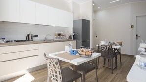A modern kitchen with white cabinets, a gas stove, and a refrigerator. There is a dining table with chairs and a small kitchen island with a sink and countertop. - MarGab Guest House Yellow Room (Napoli)