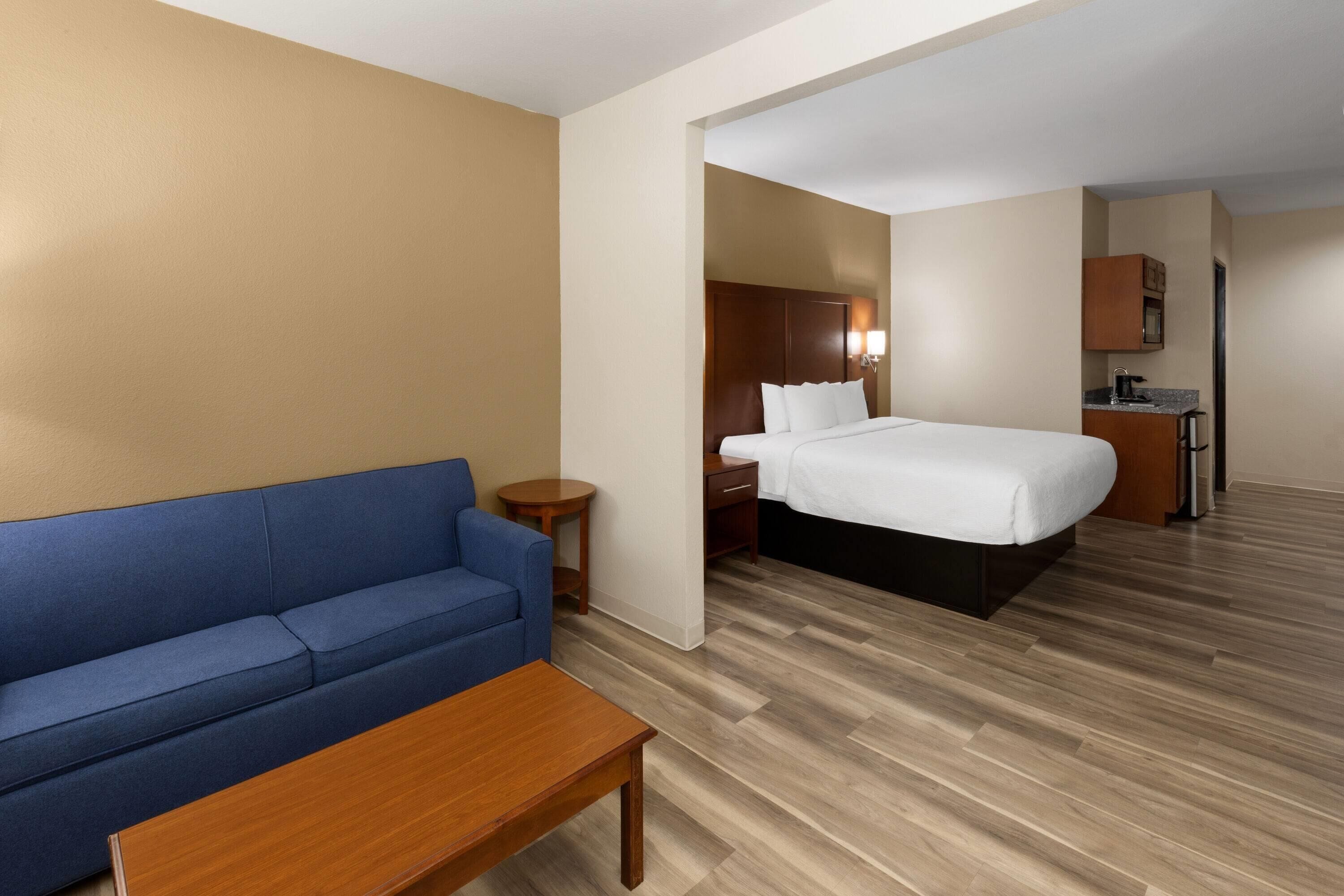 Een hotelkamer met een blauwe bank, een houten tafel, een bed met witte beddengoed, een klein rond tafeltje en een kitchenette.