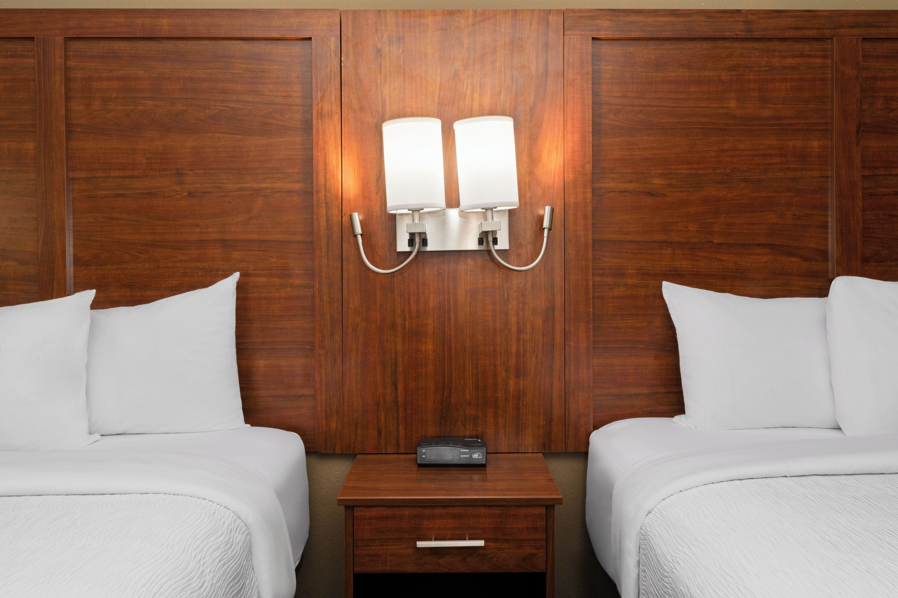 A hotel room with two beds, a wooden headboard, a nightstand with a radio, and wall-mounted lamps.