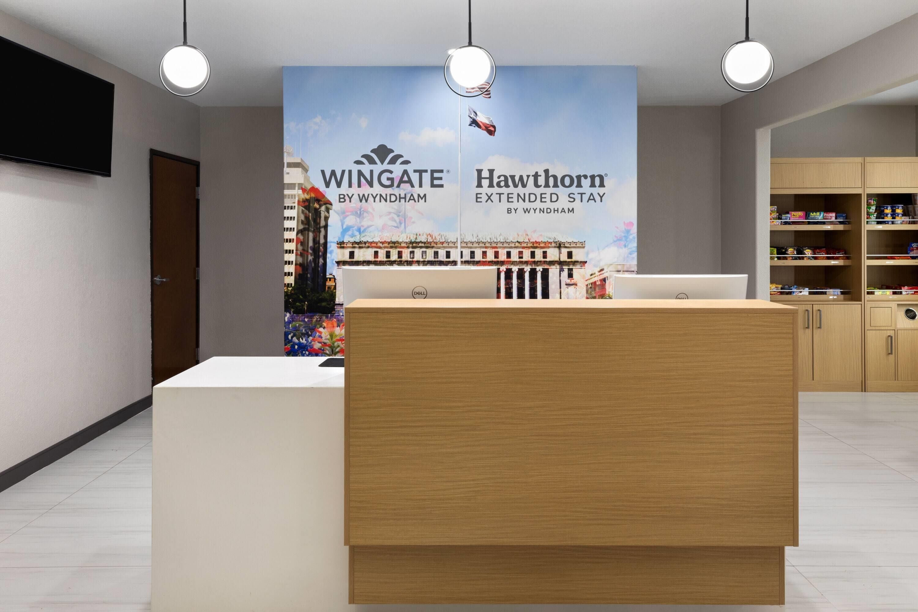 Zona de recepción de hotel con un mostrador de madera, una computadora Dell y un cartel grande que anuncia 'Wingate by Wyndham' y 'Hawthorn Extended Stay by Wyndham'.