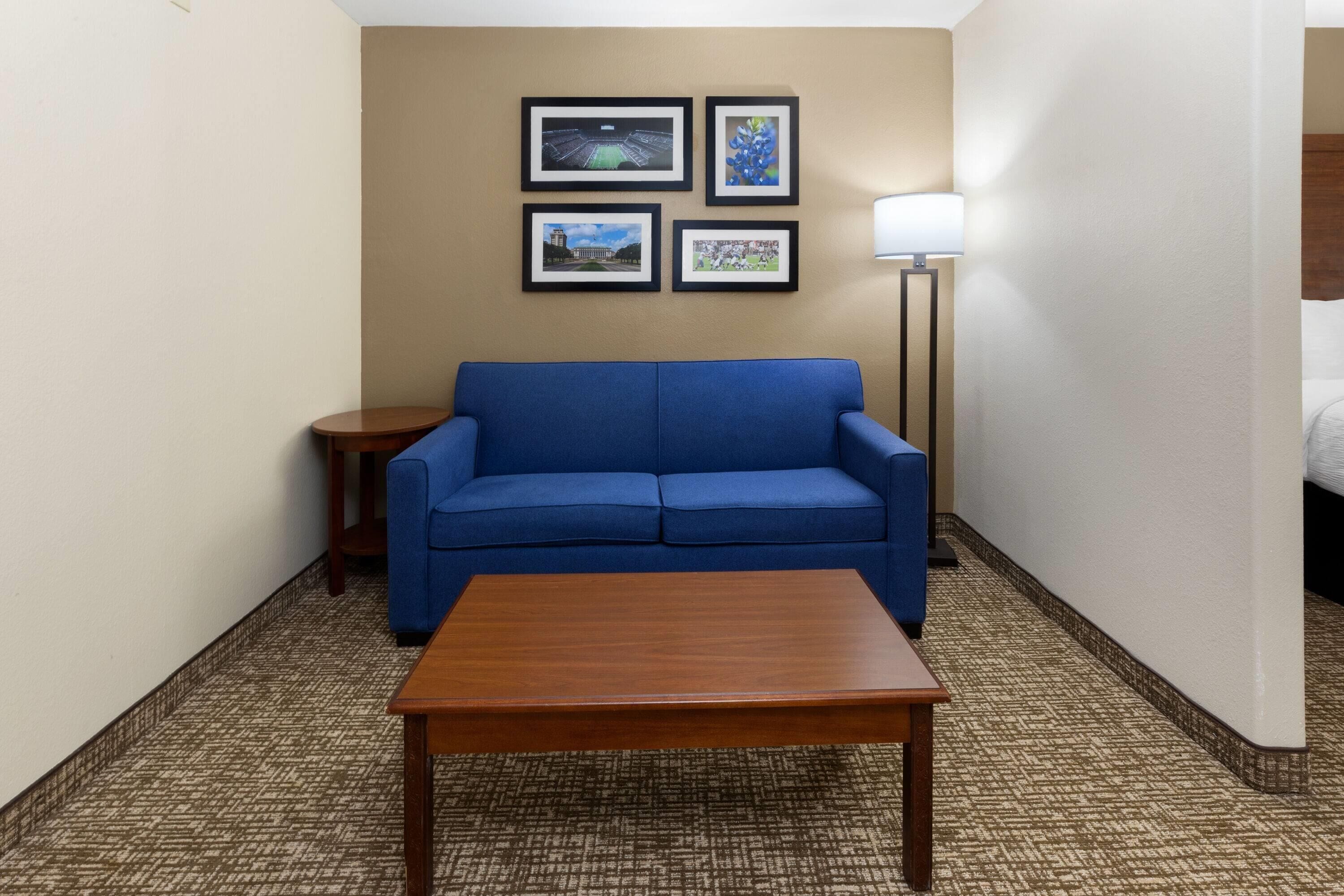A hotel room with a blue sofa, a wooden coffee table, and a lamp. There are four framed pictures on the wall.