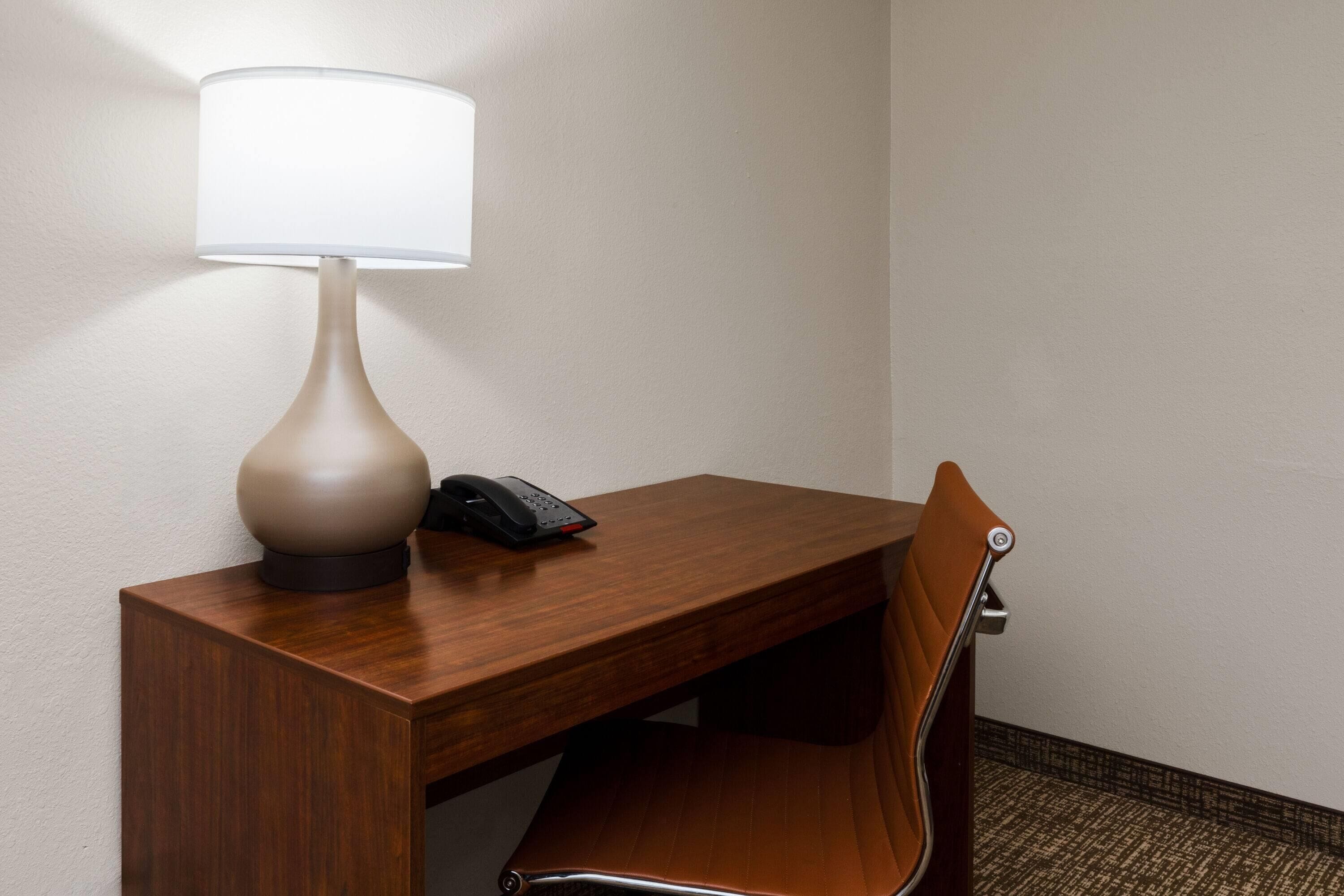 A hotel room with a wooden desk, a lamp, a telephone, and a brown chair.