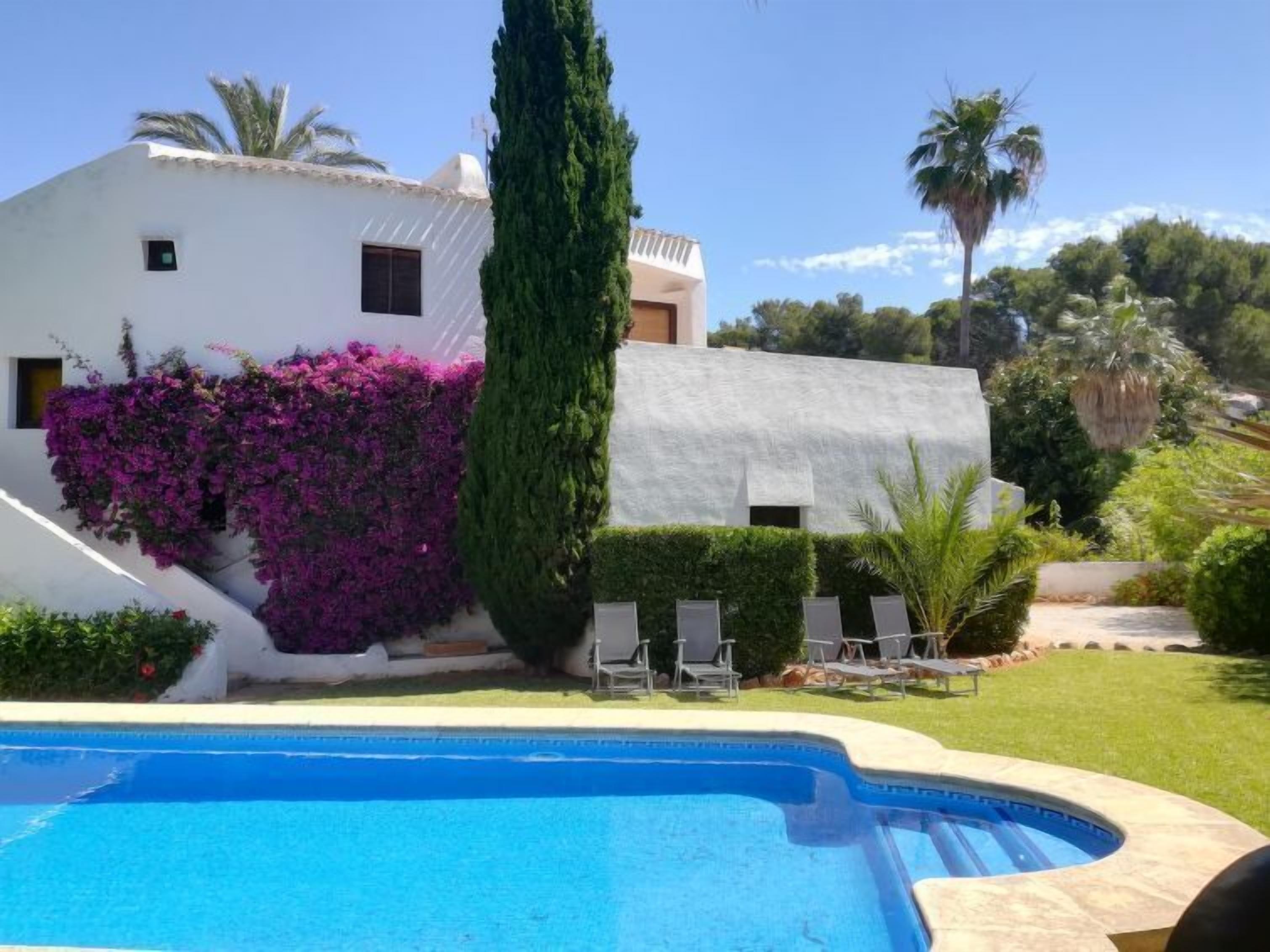 Zona de piscina amb un edifici blanc, un arbre alt i plantes de flors morades.